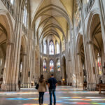 Vue intérieure réaliste de la cathédrale Saint-Étienne de Metz, montrant la nef gothique éclairée par les couleurs des vitraux et quelques visiteurs discrets.